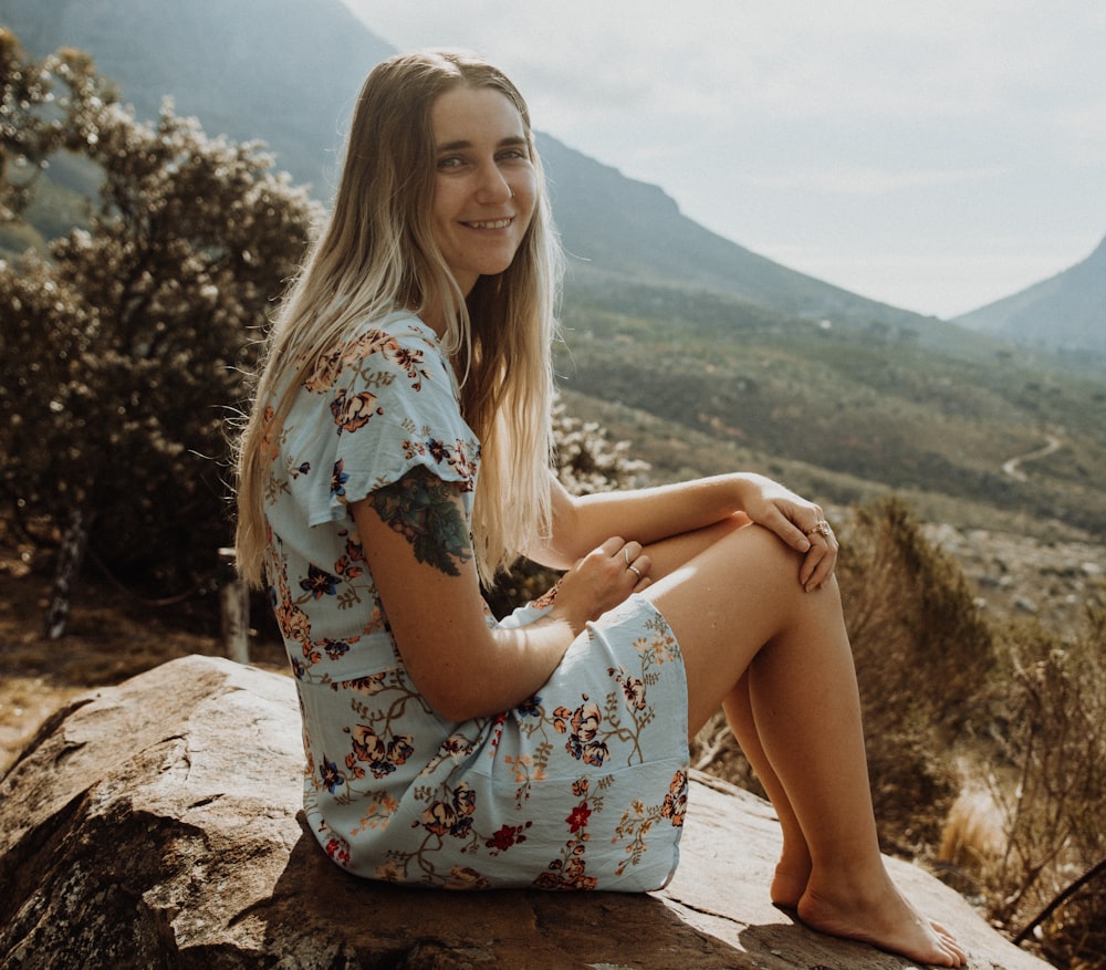 Mujer en vestido floral blanco, rojo y azul sentada en roca marrón durante el día