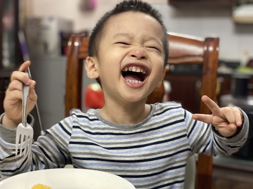 A young child with short hair is laughing joyfully, eyes closed and mouth wide open, showing teeth. The child is wearing a striped long-sleeve shirt and holding a fork in one hand while making a peace sign with the other. In the background, there is a wooden chair and blurred kitchen elements.