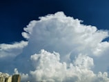 A massive cumulonimbus cloud dominates the sky, towering high above the horizon. The cloud is a bright white and has a soft, fluffy appearance, with intricate formations and textures. Below the cloud, there are several high-rise buildings, indicating an urban setting. The background sky is a deep blue, providing a contrasting backdrop to the bright cloud.