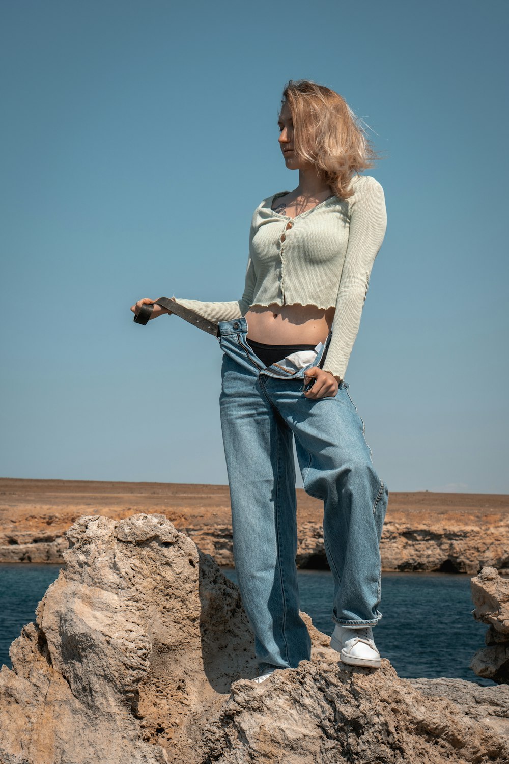 mulher na camisa branca de manga comprida e jeans jeans azuis de pé na rocha marrom perto do corpo