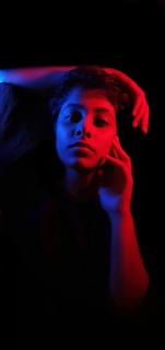 A person is lying in a relaxed pose illuminated by dramatic red and blue lighting that creates a striking contrast against the dark background. One arm is bent over their head, and the other hand is gently touching their face, creating an introspective and artistic composition.