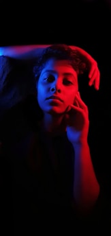 A person is lying in a relaxed pose illuminated by dramatic red and blue lighting that creates a striking contrast against the dark background. One arm is bent over their head, and the other hand is gently touching their face, creating an introspective and artistic composition.