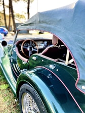 A vintage convertible car with a dark green body and brown leather interior is parked outdoors. The car has a soft top roof, wooden steering wheel, and chrome wire-spoke wheels. The background features trees and a blurry glimpse of other vehicles.