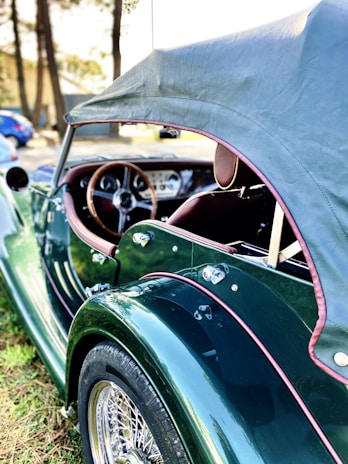 A vintage convertible car with a dark green body and brown leather interior is parked outdoors. The car has a soft top roof, wooden steering wheel, and chrome wire-spoke wheels. The background features trees and a blurry glimpse of other vehicles.