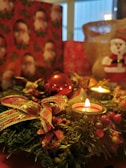 A festive arrangement featuring lit candles, a red Christmas ornament, and a decorative wreath adorned with bows and berries. In the background, holiday-themed wrapping paper with Santa Claus images and a burlap sack can be seen, contributing to a cozy and seasonal atmosphere.