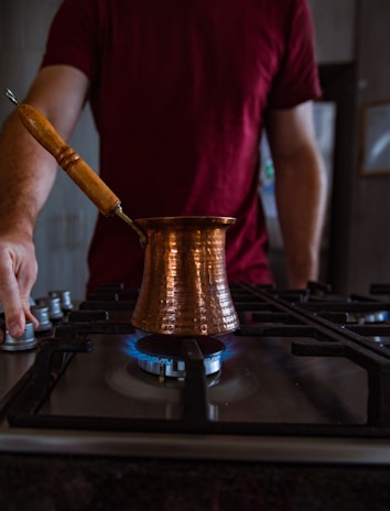 A person in a red shirt is standing by a stove, holding the knob to adjust the heat. On the stove is a traditional copper coffee pot, with a long wooden handle, placed directly on a lit burner with blue flames visible.