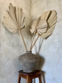 A large, textured clay pot sits on a wooden stool against a neutral-toned wall. Three dried palm leaves are arranged in the pot, creating a rustic and minimalist aesthetic.