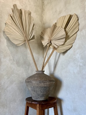 A large, textured clay pot sits on a wooden stool against a neutral-toned wall. Three dried palm leaves are arranged in the pot, creating a rustic and minimalist aesthetic.