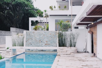 A modern outdoor area featuring a swimming pool with surrounding light stone tiles. Tall green plants are positioned alongside a stone wall, and the background displays a contemporary house with white walls and large windows. There is a bamboo roll-up shade on the patio, adding a subtle tropical element to the setting.