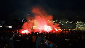 A large crowd gathers outside a stadium at night, illuminated by red flares and smoke, creating a vibrant and intense atmosphere.