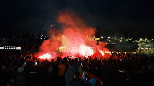 A large crowd gathers outside a stadium at night, illuminated by red flares and smoke, creating a vibrant and intense atmosphere.