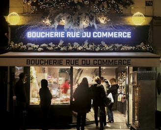 The exterior of a butcher shop, named 'Boucherie Rue du Commerce', is decorated with festive garlands and sparkling lights. Several people are gathered in front of the well-lit display window, possibly customers observing the selection inside.