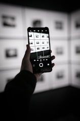 A person is holding a smartphone in their hand, capturing a photograph of a display wall with multiple black and white images arranged in a grid. The smartphone screen shows the camera interface displaying the same images in a grid layout. The environment appears to be dimly lit with contrast emphasized by the backlit display wall.