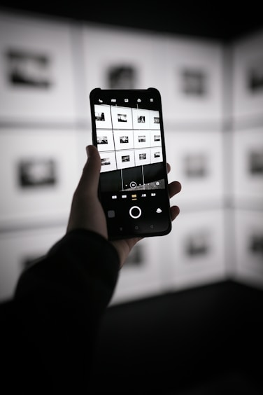 A person is holding a smartphone in their hand, capturing a photograph of a display wall with multiple black and white images arranged in a grid. The smartphone screen shows the camera interface displaying the same images in a grid layout. The environment appears to be dimly lit with contrast emphasized by the backlit display wall.