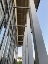 A modern architectural structure features tall, white columns supporting a metal roof with visible beams. The left side is lined with large, glass windows reflecting the clear blue sky, and small green trees can be seen in the background.