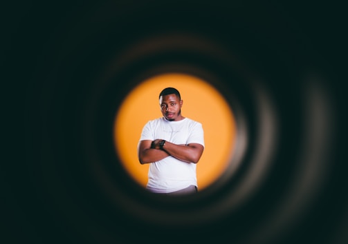 A man stands confidently with arms crossed against a bright orange background. The image is framed by a circular tunnel effect, emphasizing the subject in the center.