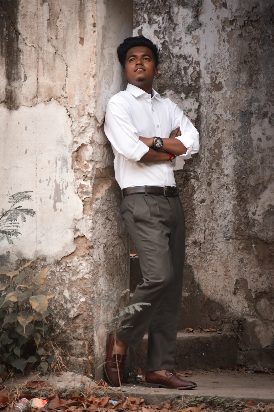 In the image, a young man is standing against a stone wall with his arms crossed and legs slightly apart. He's dressed in a crisp white shirt paired with dark pants, giving him an air of sophistication. His brown shoes add a touch of warmth to the scene. The stone wall behind him has patches of ivy growing on it, adding a natural element to the urban setting.

The image is taken from a low angle, making the man appear taller and more imposing. The background is blurred, drawing focus to the subject in the foreground.