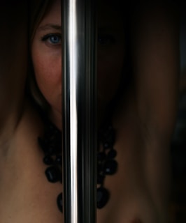 A mysterious and artistic composition featuring a person's face partially obscured by a shiny metallic pole. The image highlights one blue eye and hints of a bare shoulder adorned with a chunky black necklace, creating a striking and enigmatic visual.