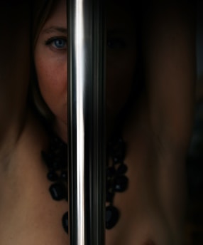 A mysterious and artistic composition featuring a person's face partially obscured by a shiny metallic pole. The image highlights one blue eye and hints of a bare shoulder adorned with a chunky black necklace, creating a striking and enigmatic visual.