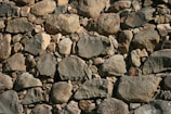 A wall constructed from an assortment of irregularly shaped stones varies in size and color. The stones are tightly packed together, displaying a range of textures and natural patterns.