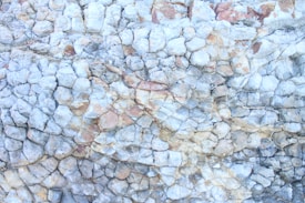 A close-up of a textured rock surface featuring irregularly shaped stones tightly packed together. The rocks display a variety of hues, predominantly in light gray and beige tones, with hints of reddish-brown. The surface shows a complex, cracked pattern, likely formed from natural geological processes.