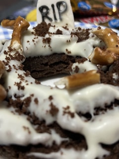 A dessert creation resembling a grave, topped with crumbled chocolate cookies, white frosting, and pretzel sticks. A small sign reads 'RIP', giving it a Halloween theme.