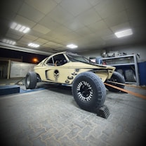 A modified off-road vehicle with large tires is parked indoors. The vehicle is beige with black detailing, including a distinctive skull logo on the side. It is positioned on a sloped platform, with straps securing it. The surrounding area appears to be a workshop with various tools and equipment visible.