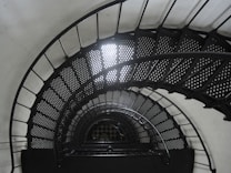 A spiral staircase with perforated metal steps winds downward in a seemingly continuous coil. The staircase is constructed with dark metal railings that create a geometric pattern as they arc and descend. Soft light filters through the upper sections, creating a contrast against the deep shadows on the stairs.