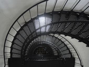 A spiral staircase with perforated metal steps winds downward in a seemingly continuous coil. The staircase is constructed with dark metal railings that create a geometric pattern as they arc and descend. Soft light filters through the upper sections, creating a contrast against the deep shadows on the stairs.