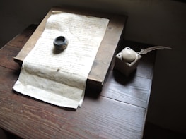 A vintage setting with a handwritten manuscript placed on a wooden desk. Beside it lies an ink pot and an old-fashioned quill pen. The lighting is soft, highlighting the weathered texture of the paper and the age of the objects.