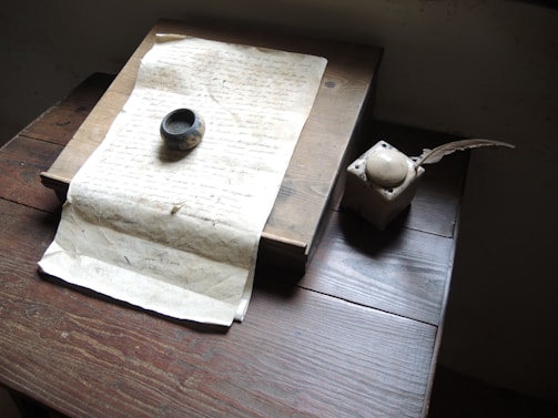 A vintage setting with a handwritten manuscript placed on a wooden desk. Beside it lies an ink pot and an old-fashioned quill pen. The lighting is soft, highlighting the weathered texture of the paper and the age of the objects.