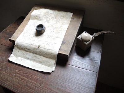 A vintage setting with a handwritten manuscript placed on a wooden desk. Beside it lies an ink pot and an old-fashioned quill pen. The lighting is soft, highlighting the weathered texture of the paper and the age of the objects.