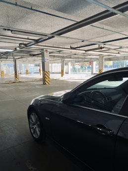 A dimly lit parking garage with a partially visible dark-colored car. The garage is mostly empty, with concrete pillars spaced evenly throughout the area. Yellow and black caution strips are visible on the pillars and signs display parking spot numbers.