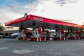 A gas station with the brand name Caltex prominently displayed on the canopy. Several fuel pumps are labeled with different numbers, situated under the red and white canopy. In the background, there are multiple parked cars and buildings, indicating it might be an area with other businesses or facilities.