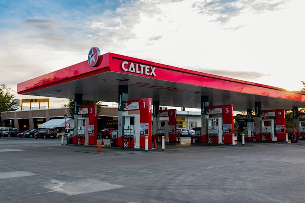 A gas station with the brand name Caltex prominently displayed on the canopy. Several fuel pumps are labeled with different numbers, situated under the red and white canopy. In the background, there are multiple parked cars and buildings, indicating it might be an area with other businesses or facilities.