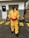 A person stands on a concrete surface outside a building, wearing a yellow protective hazmat suit with red gloves and safety goggles. Industrial pallets are visible in the background, and yellow caution lines are painted on the ground.