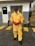 A person stands on a concrete surface outside a building, wearing a yellow protective hazmat suit with red gloves and safety goggles. Industrial pallets are visible in the background, and yellow caution lines are painted on the ground.