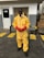 A person stands on a concrete surface outside a building, wearing a yellow protective hazmat suit with red gloves and safety goggles. Industrial pallets are visible in the background, and yellow caution lines are painted on the ground.