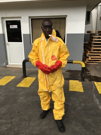 A person stands on a concrete surface outside a building, wearing a yellow protective hazmat suit with red gloves and safety goggles. Industrial pallets are visible in the background, and yellow caution lines are painted on the ground.