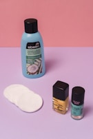 A collection of beauty products is arranged against a color-blocked background of pink and purple. There's a blue bottle of nail polish remover with a black cap and coconut scent label, several white cotton pads, a bottle of yellow nail polish, and a teal bottle of nail gel. The products are neatly positioned on a smooth surface.