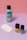 A collection of beauty products is arranged against a color-blocked background of pink and purple. There's a blue bottle of nail polish remover with a black cap and coconut scent label, several white cotton pads, a bottle of yellow nail polish, and a teal bottle of nail gel. The products are neatly positioned on a smooth surface.