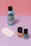 A collection of beauty products is arranged against a color-blocked background of pink and purple. There's a blue bottle of nail polish remover with a black cap and coconut scent label, several white cotton pads, a bottle of yellow nail polish, and a teal bottle of nail gel. The products are neatly positioned on a smooth surface.