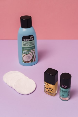 A collection of beauty products is arranged against a color-blocked background of pink and purple. There's a blue bottle of nail polish remover with a black cap and coconut scent label, several white cotton pads, a bottle of yellow nail polish, and a teal bottle of nail gel. The products are neatly positioned on a smooth surface.