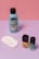A collection of beauty products is arranged against a color-blocked background of pink and purple. There's a blue bottle of nail polish remover with a black cap and coconut scent label, several white cotton pads, a bottle of yellow nail polish, and a teal bottle of nail gel. The products are neatly positioned on a smooth surface.