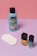 A collection of beauty products is arranged against a color-blocked background of pink and purple. There's a blue bottle of nail polish remover with a black cap and coconut scent label, several white cotton pads, a bottle of yellow nail polish, and a teal bottle of nail gel. The products are neatly positioned on a smooth surface.