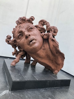 A detailed clay sculpture of a human head with flowing hair and a distressed expression, positioned on a black pedestal. The sculpture features intricate details in the facial features and textured hair, conveying a sense of motion and emotion.