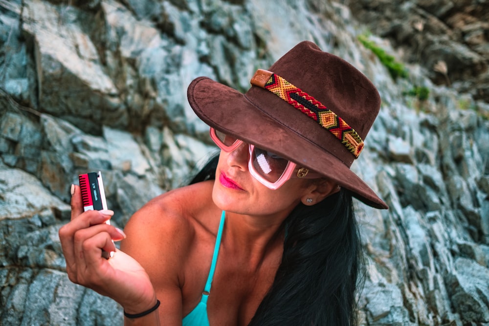 donna in top bikini blu che indossa un cappello marrone che tiene lo smartphone