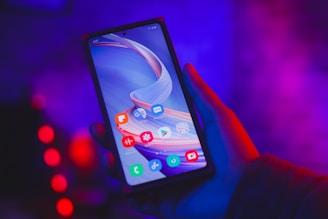 A hand holding a smartphone displaying various app icons on the screen against a vibrant, neon-lit background. The lighting creates a mix of blue and purple tones, adding a futuristic ambiance.