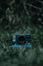 A vintage-style instant camera with a blue body and a red ring around the lens is placed on a patch of grass. The background is blurry with dark, earthy tones, contributing to a moody and mysterious atmosphere.