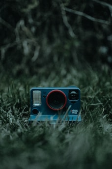 A vintage-style instant camera with a blue body and a red ring around the lens is placed on a patch of grass. The background is blurry with dark, earthy tones, contributing to a moody and mysterious atmosphere.